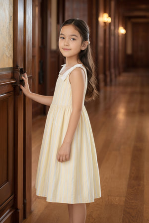 A young girl stands in a wooden hallway, wearing a yellow and white striped dress with bow shoulder details.