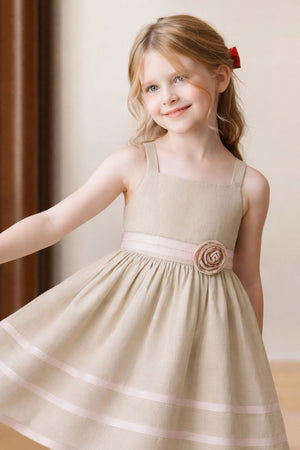 A young girl models a light sand-colored linen cami dress featuring a ribbon trim and a decorative rosette at the waist.