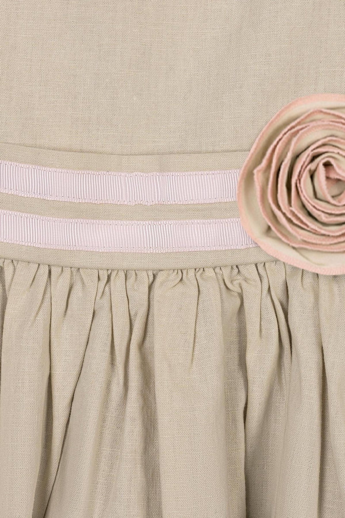 Close-up of a sand-colored dress featuring a decorative pink rosette and ribbon trim at the waist. The soft fabric showcases a gathered skirt, highlighting its elegant design for children.