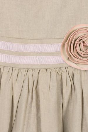 Close-up of a sand-colored dress featuring a decorative pink rosette and ribbon trim at the waist. The soft fabric showcases a gathered skirt, highlighting its elegant design for children.