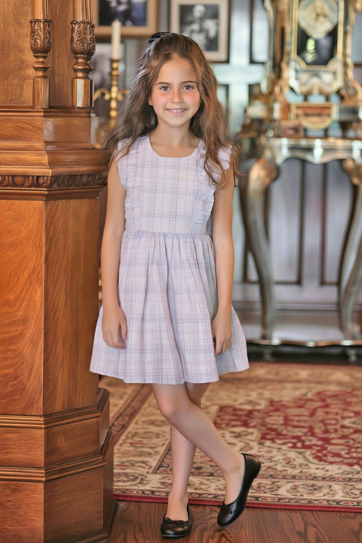 A young girl wears a light plaid dress with flutter sleeves, standing in a warmly lit room with wooden accents.