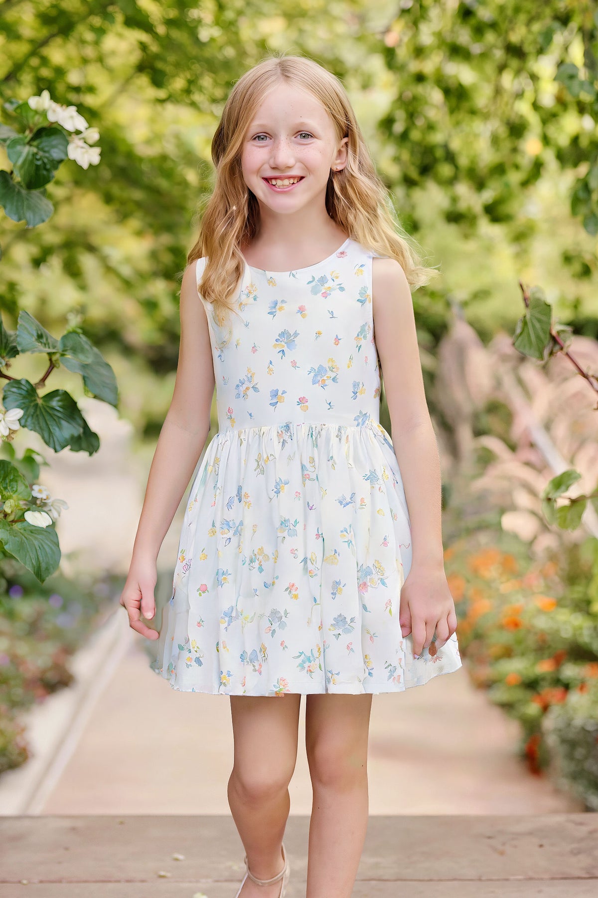 A young girl smiles while wearing a sleeveless white dress adorned with colorful floral patterns, standing in a lush garden.