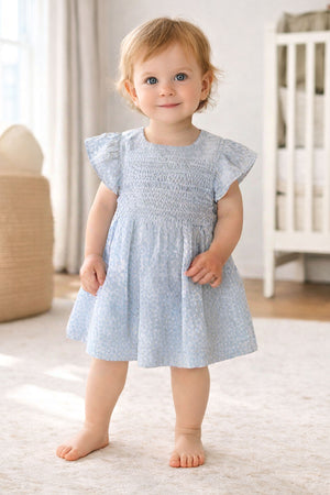 A baby girl wearing a blue smocked puff sleeve dress stands barefoot on a soft carpet in a nursery.