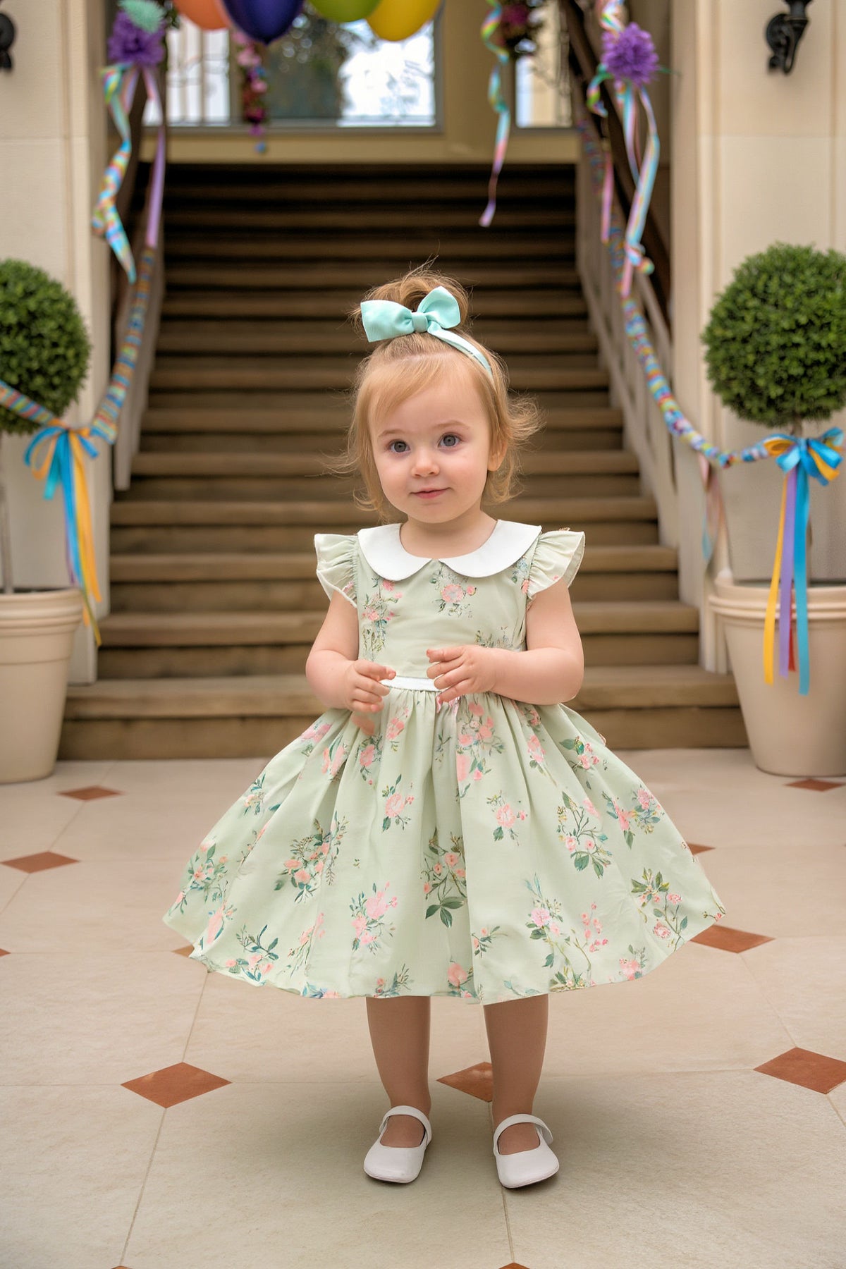 A toddler stands on a tiled floor, wearing a mint green floral dress with a Peter Pan collar and matching.
