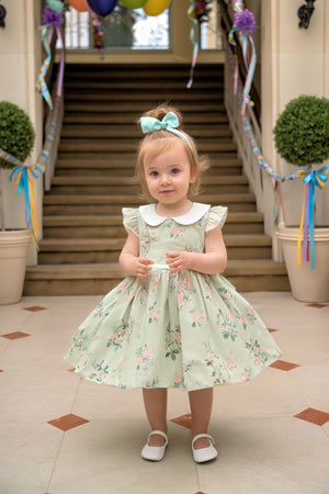 A toddler stands on a tiled floor, wearing a mint green floral dress with a Peter Pan collar and matching.