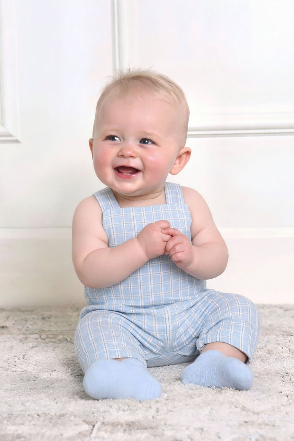A smiling baby sits on a soft surface, wearing a light blue checkered overall romper and matching blue socks.