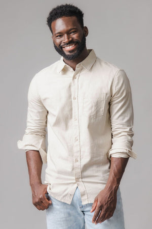 A smiling man wears a light sand-colored linen button-down shirt, featuring a chest pocket and rolled-up sleeves, paired with light blue jeans against a neutral gray background.