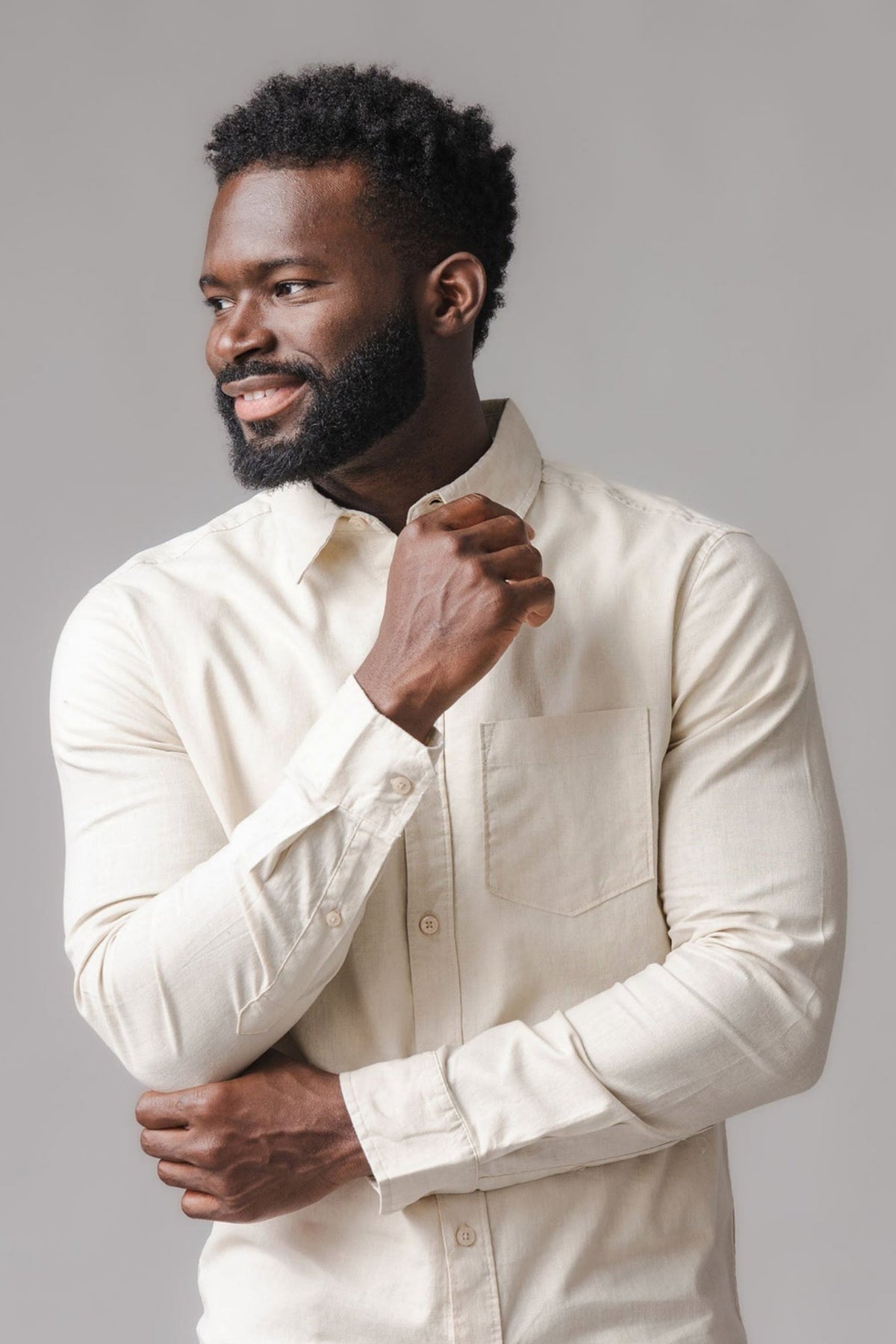 A man wearing a light sand-colored linen button-down shirt stands against a neutral background. The shirt features a chest pocket and a classic long sleeve design, highlighting its casual style.