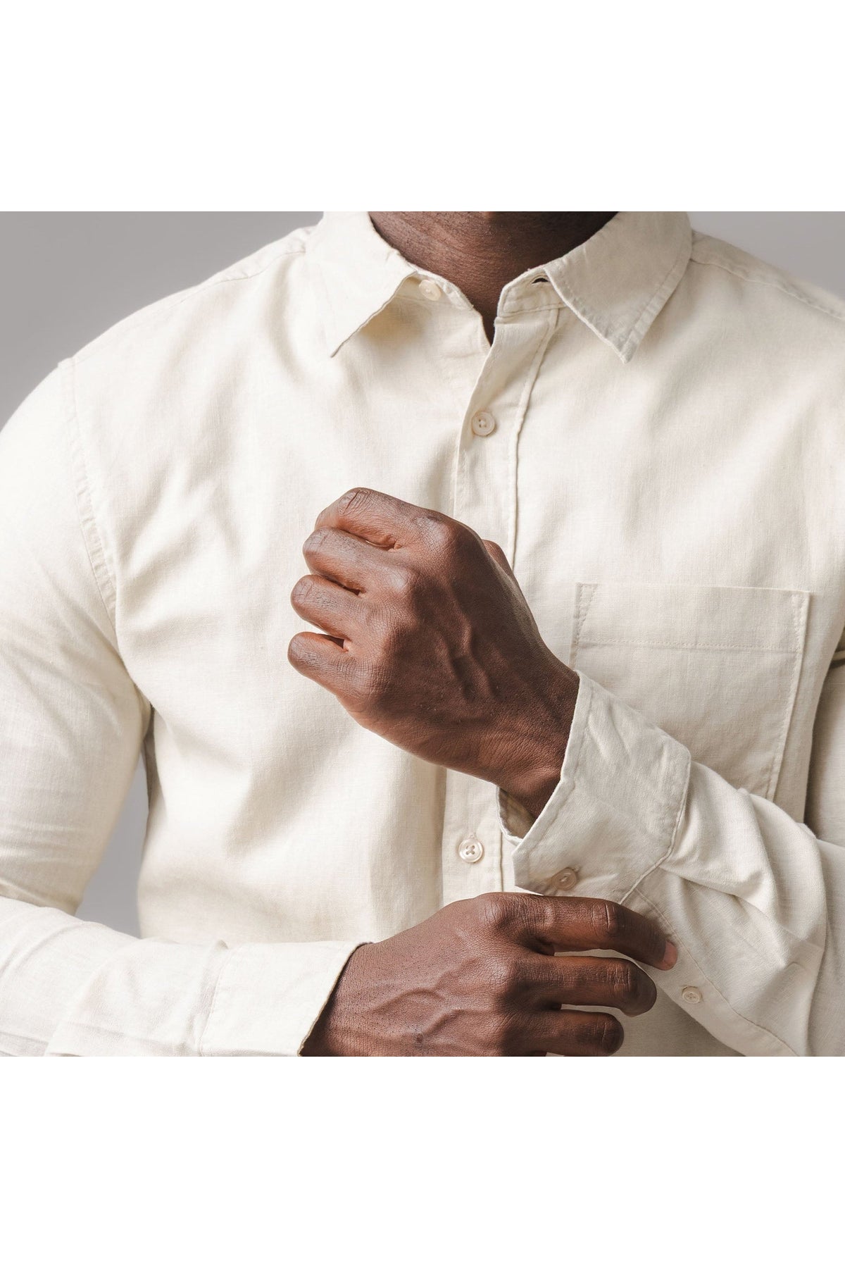A model is adjusting the cuff of a light sand-colored linen button-down shirt, featuring a functional chest pocket and classic collar, showcasing its casual yet refined style.