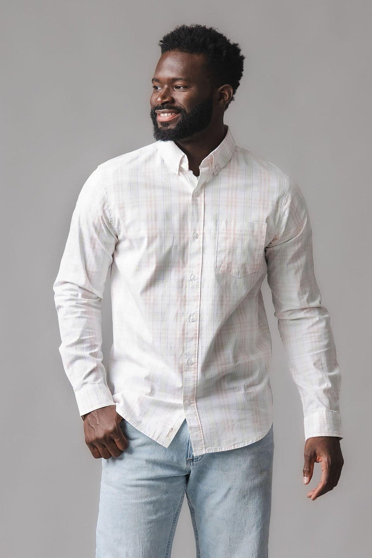 A man smiles while wearing a long-sleeve, button-down shirt in a soft, light-colored Chateau plaid pattern. The shirt features a functional chest pocket and is made from organic cotton.