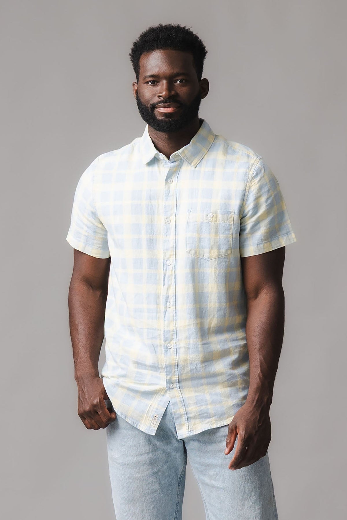A man stands confidently wearing a short sleeve button-down shirt featuring a sunlit plaid pattern in soft yellow and blue hues.