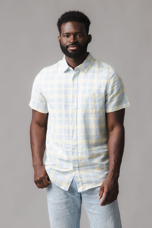 A man stands confidently wearing a short sleeve button-down shirt featuring a sunlit plaid pattern in soft yellow and blue hues.