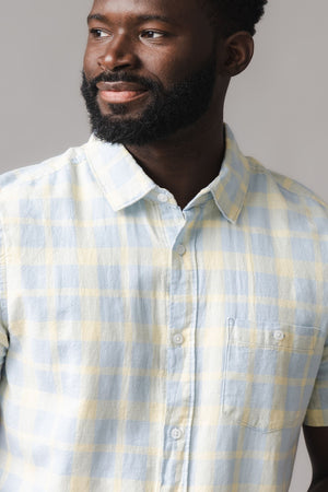 A man wearing a short-sleeve button-down shirt in a sunlit plaid pattern, featuring soft linen and organic cotton fabric. The shirt has a functional chest pocket and a relaxed fit.