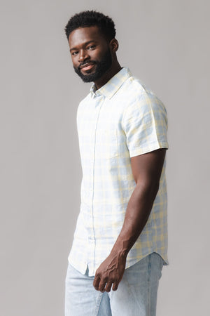 A man poses in a soft, short-sleeve button-down shirt featuring a sunlit plaid pattern in light yellow and blue. The shirt is made from a linen and organic cotton blend.