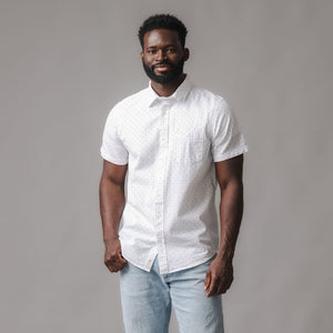 A man stands confidently wearing a white short sleeve button-down shirt with a subtle dot pattern. The shirt features a chest pocket and is crafted from organic cotton for comfort.