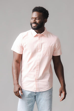 A man is wearing a light pink, short sleeve shirt made from soft organic cotton. He smiles while standing against a neutral gray background, showcasing a casual and comfortable style.