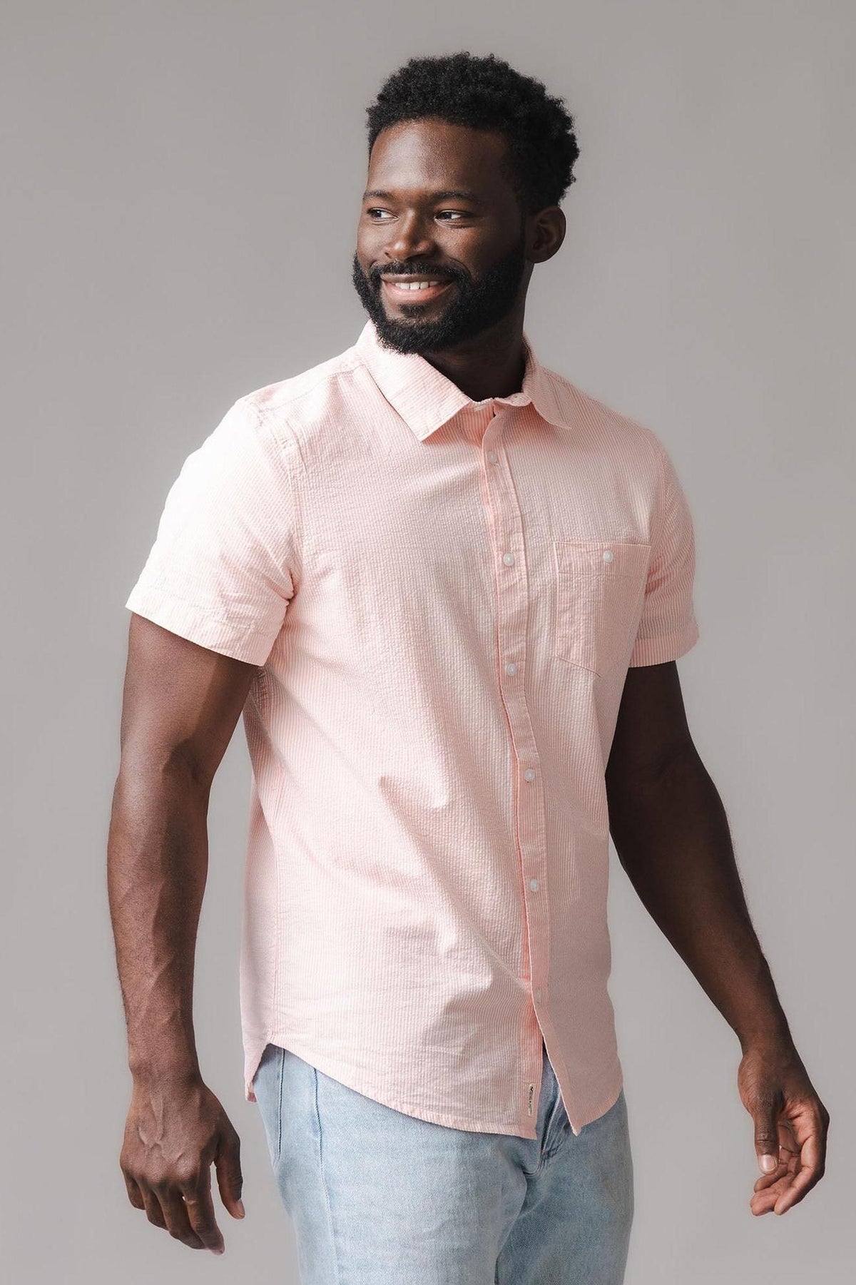 A man is wearing a light pink short-sleeve shirt with a button-down collar, featuring a subtle seersucker texture. The shirt is made from organic cotton, offering a relaxed, stylish look.