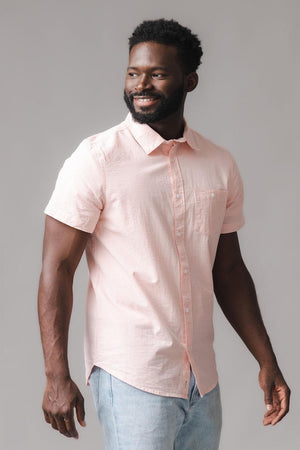 A man is wearing a light pink short-sleeve shirt with a button-down collar, featuring a subtle seersucker texture. The shirt is made from organic cotton, offering a relaxed, stylish look.