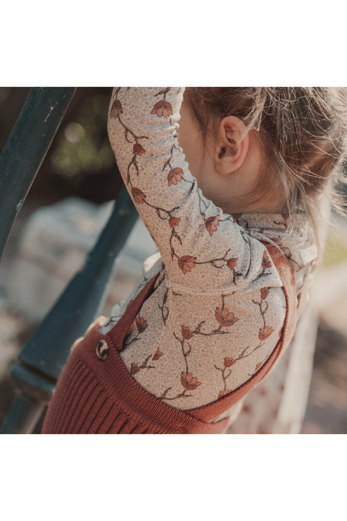 A child wears a soft, patterned turtleneck featuring a floral design, paired with a cozy rust-colored overall. The child is playfully climbing a staircase in a natural outdoor setting.