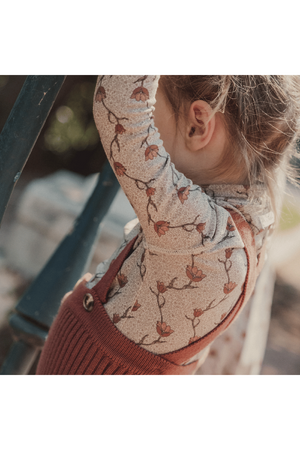 A child wears a soft, patterned turtleneck featuring a floral design, paired with a cozy rust-colored overall. The child is playfully climbing a staircase in a natural outdoor setting.