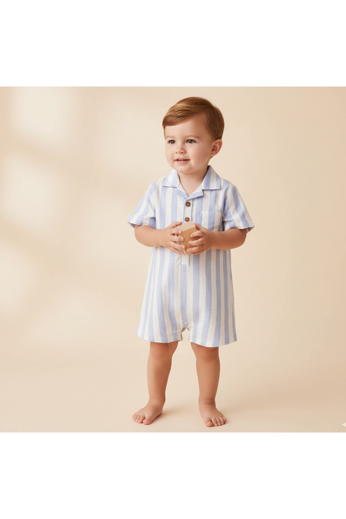 A young child stands barefoot, wearing a blue and white striped organic collared romper. The lightweight fabric features a smart collar and wooden buttons, perfect for warm, sunny days.