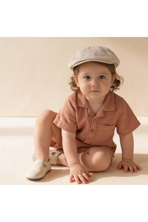 A young child sits on a light surface, wearing a clay-colored organic collared romper with buttons and a pocket.