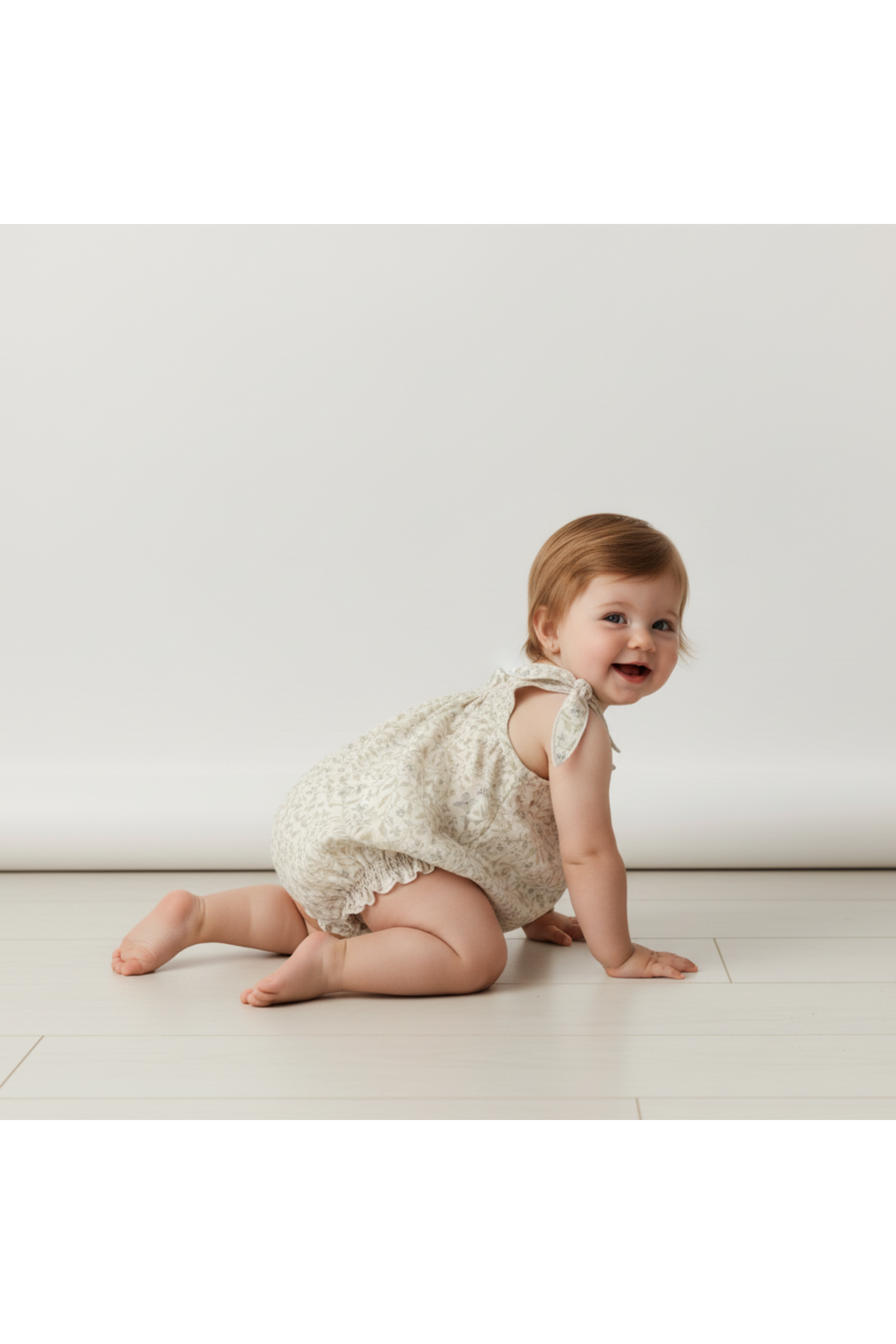 A toddler crawls on a light wooden floor, wearing a soft, organic cotton bubble romper with tie shoulder straps in a delicate cream print, showcasing a relaxed and comfortable fit.