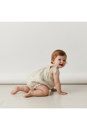 A toddler crawls on a light wooden floor, wearing a soft, organic cotton bubble romper with tie shoulder straps in a delicate cream print, showcasing a relaxed and comfortable fit.