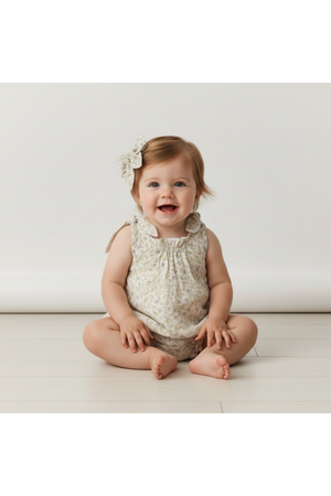 A smiling baby sits on a light wooden floor, wearing a floral-patterned romper. She has a matching organic cotton hair bow, complementing her playful and cheerful demeanor.