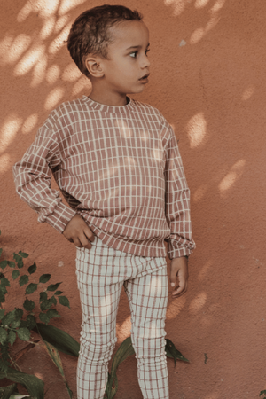 A young child stands against a warm, earthy wall, wearing a mauve check printed cotton sweatshirt paired with coordinating checkered leggings, showcasing a cozy and stylish outfit suitable for toddlers.