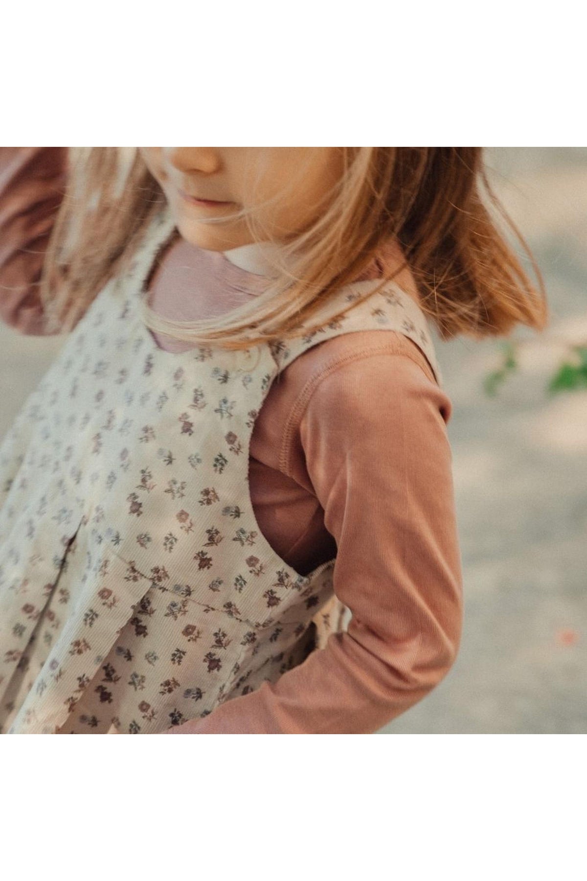 A young girl is wearing a floral corduroy dress over a long-sleeve top, showcasing a soft color palette.