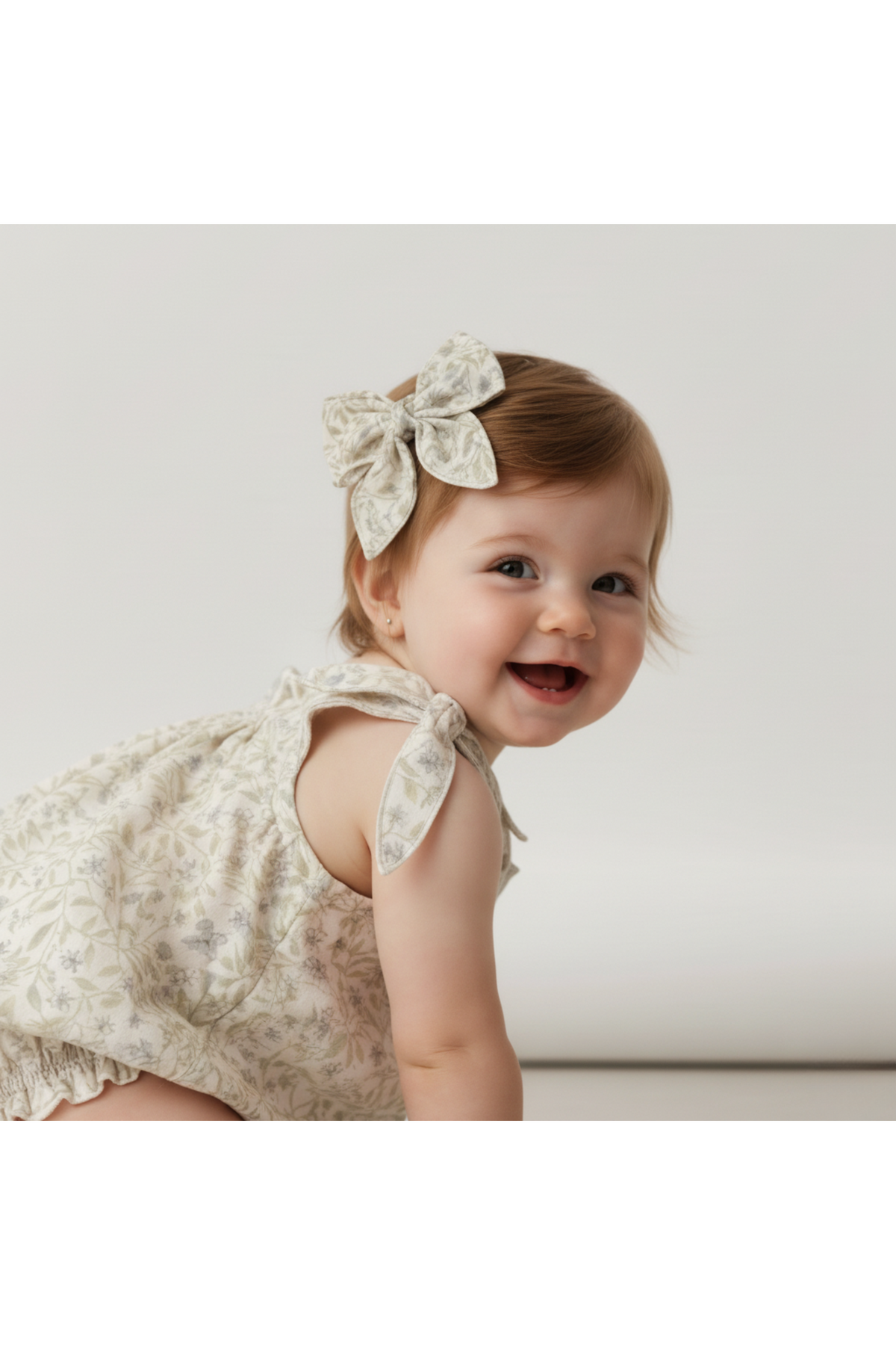 A smiling baby wearing a light floral-patterned outfit and a large bow clip in their hair sits on a neutral background, showcasing the charm and comfort of organic cotton materials.