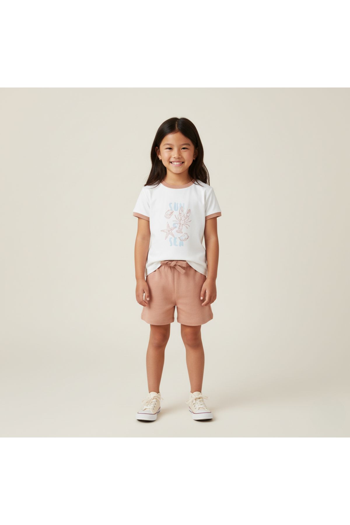 A young girl smiles, wearing a soft white graphic tee featuring a beach design and light pink shorts.