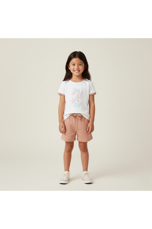 A young girl smiles, wearing a soft white graphic tee featuring a beach design and light pink shorts.