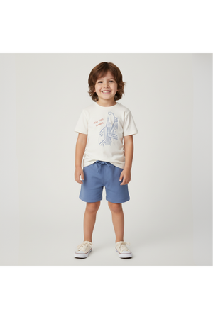 A young child stands smiling, wearing a cream graphic tee featuring a blue New York illustration and light blue shorts.