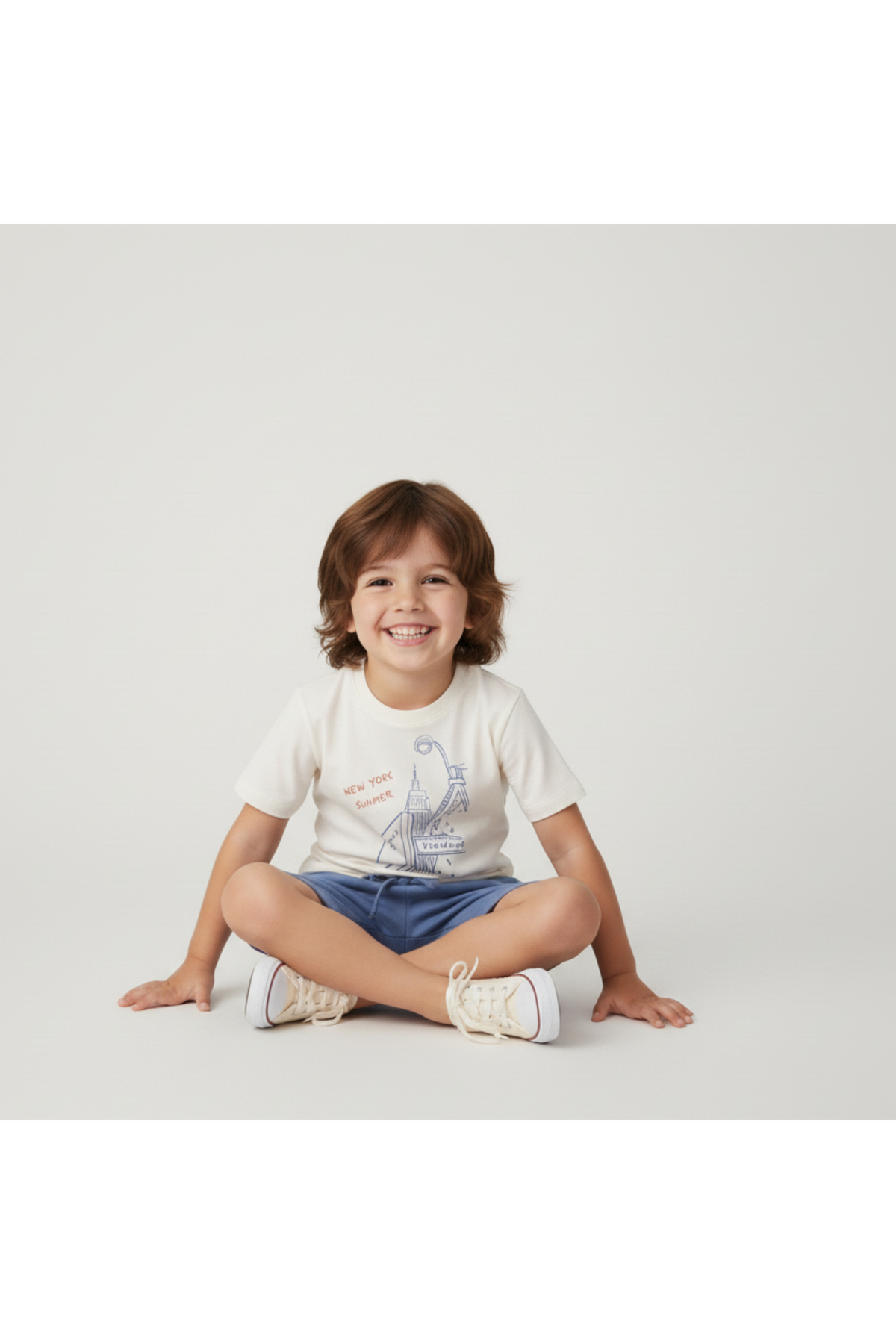 A smiling child sits cross-legged, wearing a cream organic cotton graphic tee featuring a New York print and blue shorts. The neutral background enhances the playful and comfortable outfit.