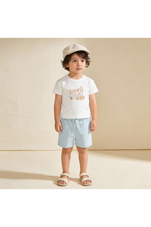 A young child stands in a light setting, wearing a white graphic tee with a vintage truck design and soft blue shorts.