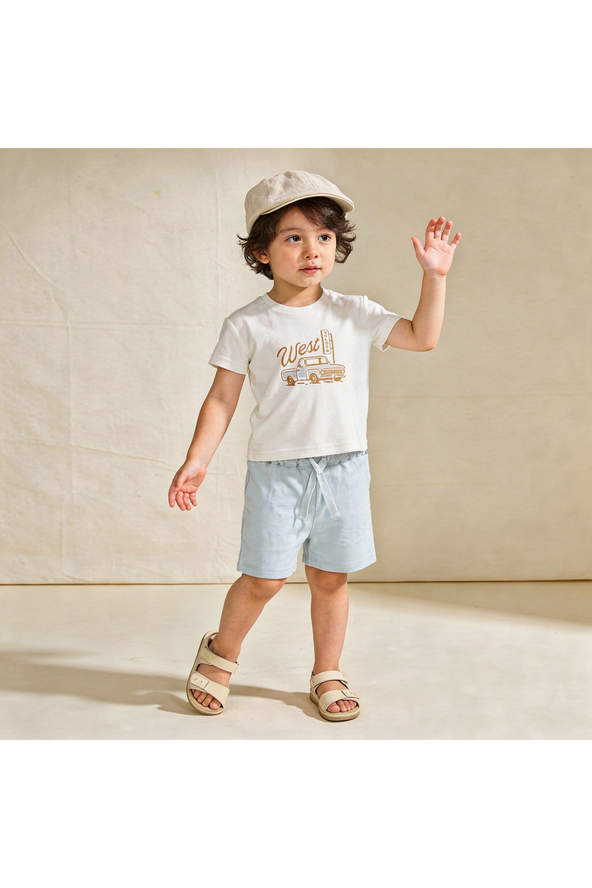 A young child in a white graphic tee with a West print and light blue shorts stands against a neutral background.