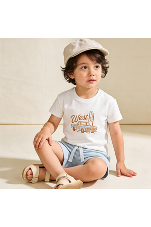 A young child sits on a light-colored surface, wearing a white graphic tee with a West print and light blue shorts.