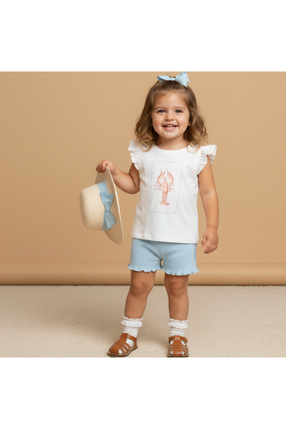 A smiling young girl wears a sleeveless white top featuring a lobster print and blue shorts, holding a straw hat with a blue bow.