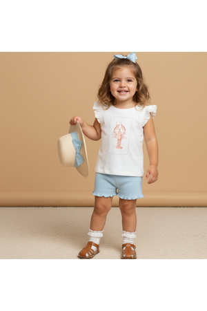 A smiling young girl wears a sleeveless white top featuring a lobster print and blue shorts, holding a straw hat with a blue bow.