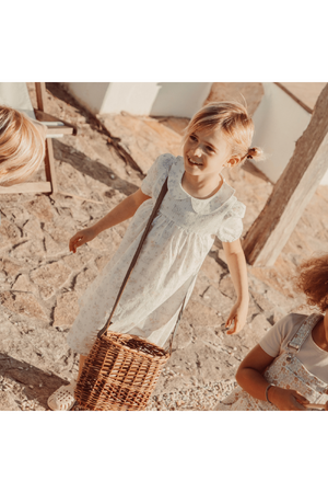 A young girl smiles while wearing a light blue dress adorned with small floral patterns. She holds a woven basket, standing on a stone pathway in a sunlit outdoor setting.