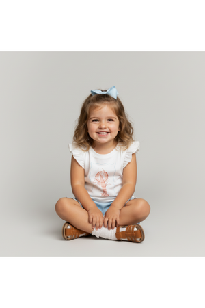 A smiling young girl sits cross-legged, wearing a sleeveless white top with a lobster design and light blue shorts.