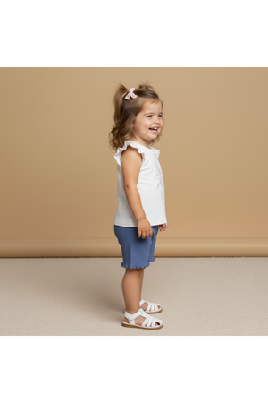 A smiling toddler stands sideways, wearing a white sleeveless top with ruffled edges and blue shorts. The outfit is made from organic cotton, perfect for warm weather and playful activities.