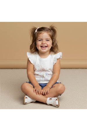 A smiling toddler sits cross-legged on a soft surface, wearing a white sleeveless top with flutter sleeves and a floral design, paired with denim shorts and sandals.