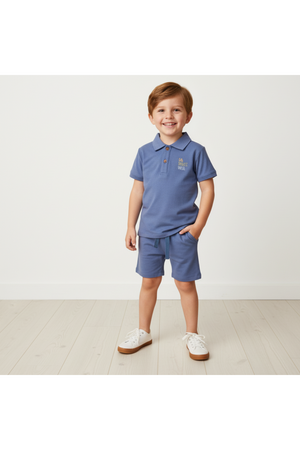 A young boy stands on a light wooden floor, wearing a slate-colored organic cotton polo shirt and matching shorts.