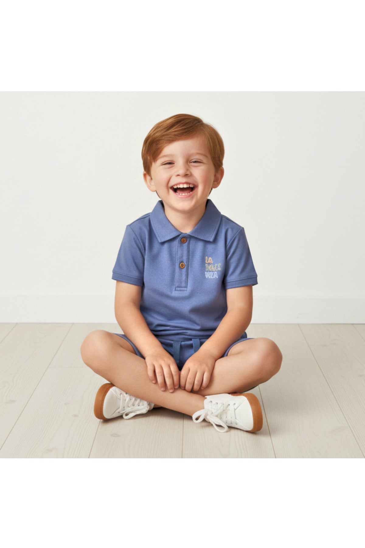 A smiling child sits cross-legged on a light wooden floor, wearing a soft slate blue polo shirt and matching shorts.