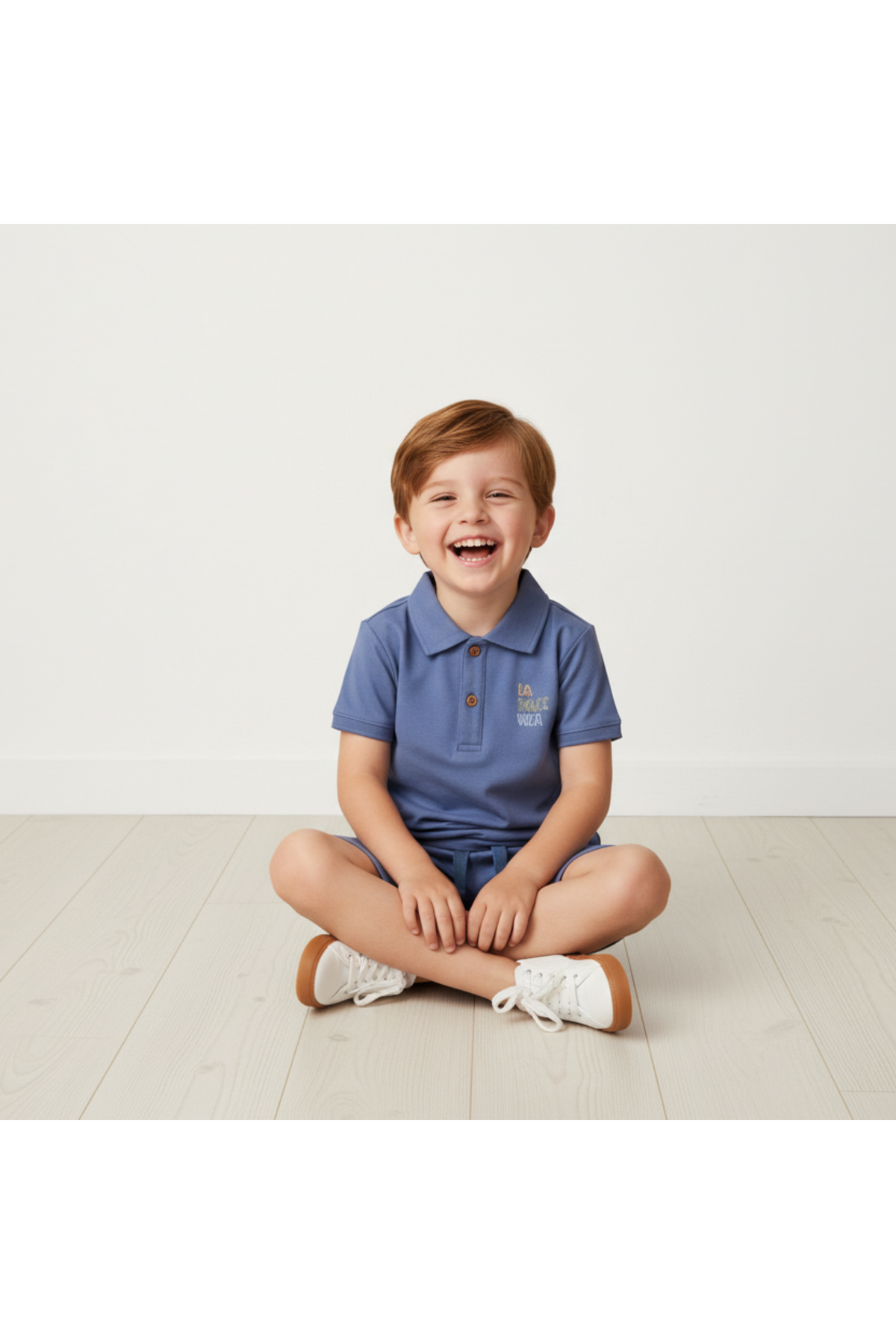 A smiling child sits on a light wooden floor, wearing a slate blue organic polo shirt and matching shorts. The outfit is comfortable and suitable for casual playdates.