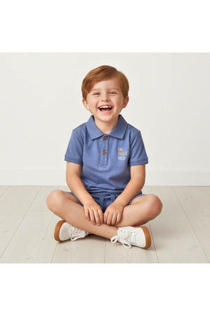 A smiling child sits cross-legged on a light wooden floor, wearing a soft slate blue polo shirt and matching shorts.