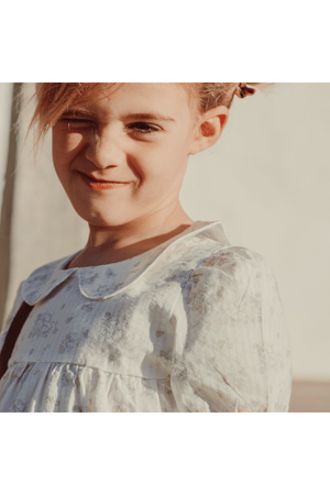 A young girl smiles playfully while wearing a light, floral-patterned dress with a classic collar. The dress features short puff sleeves and a soft, airy fabric, perfect for special occasions.